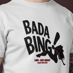 Può includere: T-shirt bianca con le parole "Bada Bing" in grandi lettere nere in grassetto. Sotto il testo c'è una grafica nera di una donna con un accento rosso. Il testo "Lodi - New Jersey" è sotto la grafica.