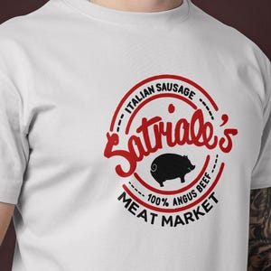 Könnte beinhalten: Weißes T-Shirt mit einem roten und schwarzen kreisförmigen Logo. Das Logo lautet "Satriale's Italian Sausage Meat Market" und zeigt eine Schweineillustration. Der Text enthält auch "100% Angus Beef."