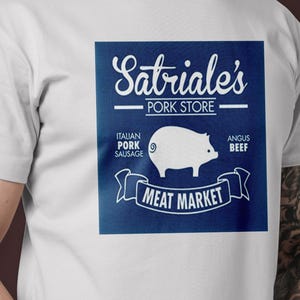 Könnte beinhalten: Weißes T-Shirt mit einem blau-weißen Logo, auf dem "Satriale's Pork Store" und "Meat Market" steht. Das Logo zeigt außerdem ein Schwein und den Text "Italian Pork Sausage" und "Angus Beef".