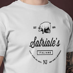 Könnte beinhalten: Weißes T-Shirt mit einer schwarzen Grafik eines Schweins über dem Text "Satriale's Italiano" und anderem Text. Das Shirt enthält den Text "EST 1960" und "Pork Store Deli Meats, NJ, Good Food & Espresso".