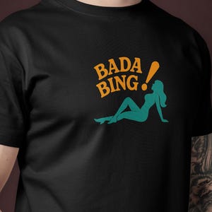 Può includere: T-shirt nera con le parole "Bada Bing!" in arancione e una silhouette turchese di una donna reclinata. La maglietta è realizzata in materiale morbido e ha maniche corte.