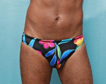 May include: A man wearing black swim trunks with a floral print. The print features bright pink, yellow, blue, and green flowers and leaves on a black background.