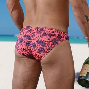 May include: A man wearing pink and black floral print swimming trunks stands on a white sandy beach holding a bottle of champagne.