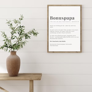 Könnte beinhalten: Ein gerahmter Druck mit dem deutschen Wort "Bonuspapa" und seiner Definition. Der Druck befindet sich in einem Holzrahmen. Eine braune Vase mit grünen Zweigen steht auf einem Holztisch. Der Druck ist an einer weißen Wand.