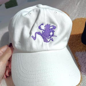 May include: A white baseball cap featuring a purple embroidered graphic of a stylized running figure on the front panel. The cap has a structured crown, a curved brim with stitched detailing, and a small button at the top center of the crown.