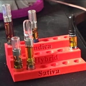 May include: A red tiered organiser holds several vape cartridges. The cartridges are clear with various amber-coloured oils. The organiser has the words "Indica", "Hybrid", and "Sativa" printed on it.
