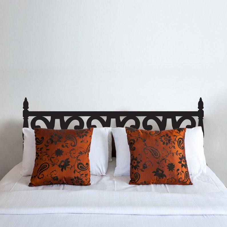 Puede incluir: Un adhesivo de cabecero de metal negro con un dise&ntilde;o decorativo de desplazamiento. El adhesivo se aplica a una pared blanca sobre una cama con ropa de cama blanca y dos almohadas de tiro estampadas.