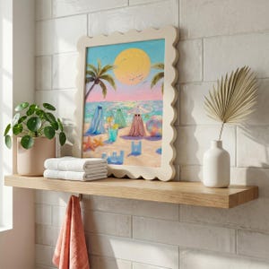 Tiny Ghosts Beach Day Wall Art displayed in a unique wavy cream frame on a wooden bathroom shelf next to a green plant and white towels. The playful pastel colors of the summer ghosts provide a cheerful contrast against the clean white tile wall in this bright maximalist interior. An ideal example of quirky bathroom art that transforms a functional space into a whimsical summer retreat... — bring warmth home.