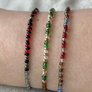 Peut inclure: Trois bracelets en perles sur un poignet. Les bracelets sont faits de petites perles colorées en rouge, noir, vert, or, argent et bleu.
