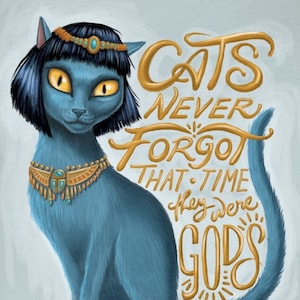 May include: A blue cat with a gold crown and necklace sits against a light blue background. The cat has yellow eyes and a black bob. The text "Cats never forgot that time they were gods" is written in gold cursive lettering.