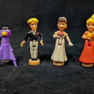 May include: Four plastic figurines, two brides and two grooms, with a purple stand. The brides are wearing white dresses and holding bouquets. The grooms are wearing black suits and holding buttonholes. The stand is a purple rocket ship with a clear top.