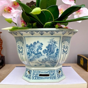 Pode incluir: Um vaso de porcelana azul e branco com um desenho de paisagem, segurando orquídeas rosa e brancas. O vaso tem uma base hexagonal e uma borda detalhada. As orquídeas têm folhas verdes e pétalas rosa delicadas.