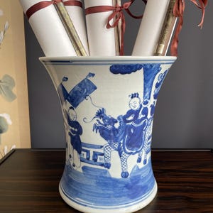 Peut inclure: Un vase en céramique blanche orné d'illustrations peintes à la main en bleu représentant des personnages et un dragon. Le vase présente un bord évasé et contient plusieurs rouleaux de papier attachés par des rubans marron fins.