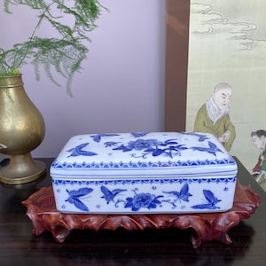 Peut inclure: Boîte rectangulaire en porcelaine blanche ornée de motifs floraux et de papillons bleus. La boîte est posée sur un support en bois foncé. Le couvercle présente un motif floral central et une bordure bleue et blanche.