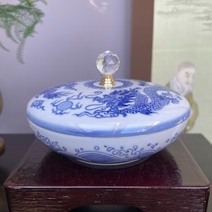 Könnte beinhalten: Weiß-blaue Keramik-Schmuckschatulle mit Deckel mit Drachendesign. Der Deckel hat einen Kristallknauf mit goldfarbenem Sockel. Die Schatulle steht auf einem dunklen Holzständer.