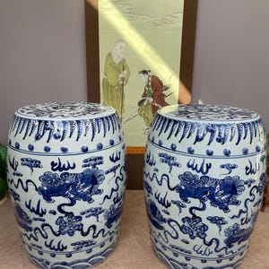 Può includere: Due sgabelli da giardino in ceramica blu e bianca abbinati. Gli sgabelli hanno una forma cilindrica con un elaborato disegno di draghi e nuvole. Lo sfondo presenta un dipinto asiatico tradizionale.