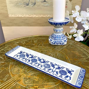 Könnte beinhalten: Ein weißes rechteckiges Keramikserviertablett mit blauem Blumenmuster und blauem Rand. Eine weiße Kerze steht auf einem blau-weißen Kerzenständer aus Keramik. Der Hintergrund umfasst ein Gemälde und weiße Blumen.