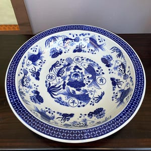 Könnte beinhalten: Eine weiße Keramik-Servierschüssel mit blauem Fisch- und Blumendesign. Die Schüssel hat einen breiten Rand mit einem geometrischen Muster. Das Innere zeigt Kois, Lotusblumen und chinesische Schriftzeichen.