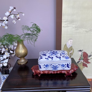 Puede incluir: Una caja de porcelana blanca y azul con un diseño floral descansa sobre un soporte de madera tallada. La caja está sobre una mesa de madera oscura, con un jarrón dorado y un arreglo floral en el fondo.