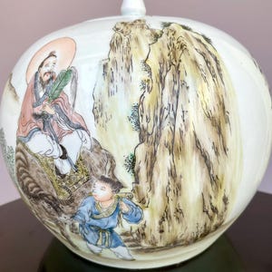 Könnte beinhalten: Ein weißes Keramik-Ingwerglas mit Deckel, das eine handbemalte Szene zeigt: eine Figur mit Heiligenschein auf einem großen Tier, begleitet von einer Person in blauer Kleidung, vor einer Kulisse aus hohen, strukturierten Felsen in Brauntönen.