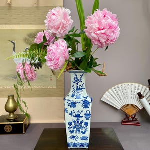 Op de afbeelding: Een hoge, vierkante keramische vaas met een witte basis en ingewikkelde blauwe bloem- en decoratieve patronen. De vaas is gevuld met een groot arrangement van levendige roze pioenrozen en lange groene bladeren voor een gebalanceerd geheel.