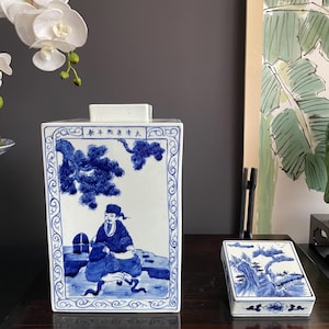 Puede incluir: Un tarro rectangular de porcelana blanca con escenas pintadas a mano en azul bajo vidriado que muestran una figura sentada bajo un pino y una tapa rectangular a juego. El tarro presenta caracteres chinos y un patrón de borde decorativo.