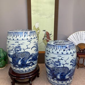 Puede incluir: Dos taburetes de jardín de cerámica azul y blanca con motivos de dragones. Los taburetes cilíndricos presentan un diseño detallado de dragón, olas y follaje. Se apoyan en soportes de madera oscura, mostrando arte asiático tradicional.