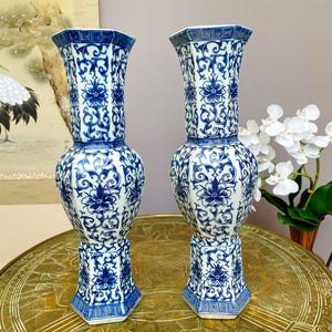 May include: A pair of tall, hexagonal ceramic vases featuring a white base with intricate blue floral and scrolling vine patterns. Each vase has a flared rim, a bulbous midsection, and a geometric base, all decorated with consistent blue motifs.