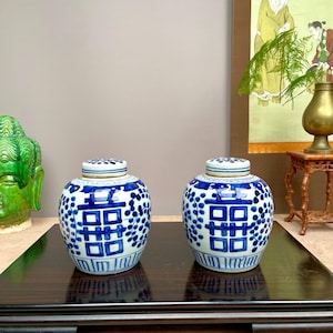 Puede incluir: Dos tarros de jengibre de cerámica azul y blanca con tapas. Los tarros presentan un patrón repetitivo de puntos azules y un diseño cuadrado azul central. Los tarros se exhiben sobre una superficie de madera oscura.