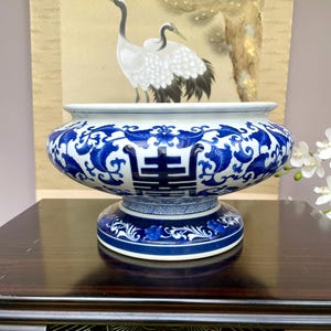 Puede incluir: Un cuenco de porcelana azul y blanco con un cuerpo ancho y redondeado y una base de pedestal. El cuenco presenta intrincados patrones florales y geométricos, con un carácter chino central. El fondo incluye una pintura de grullas.