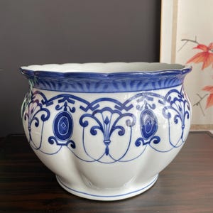 Op de afbeelding: Een witte keramische plantenpot met een geschulpte rand en een afgerond, gegroefd lichaam. De buitenkant is voorzien van een blauw geschilderd patroon van herhalende ovale medaillons en gestileerde bloemmotieven met een blauwe rand.