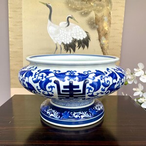 Könnte beinhalten: Eine runde Porzellanschale mit weißem Untergrund, verziert mit komplexen blauen floralen Rankenmustern und einem zentralen chinesischen Schriftzeichen. Die Schale hat einen breiten, ausgestellten Rand und steht auf einem runden Sockel.