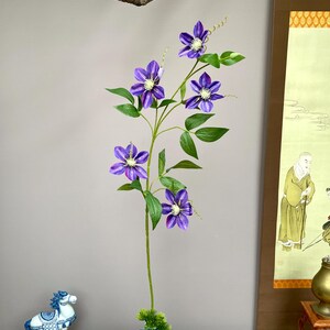May include: A tall artificial stem featuring five vibrant purple clematis flowers with white centres, green leaves, and thin curling tendrils. The stem is arranged in a round, blue and white patterned ceramic vase with a small patch of green moss.