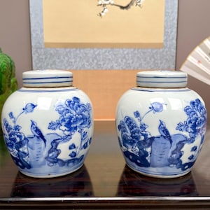 May include: Two matching white and blue ceramic jars with lids. Each jar features a floral and bird design in blue. The jars are round with a small, flat lid. The background includes a painting and a fan.