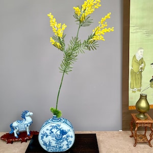 May include: A blue and white ceramic vase with a floral design holds a sprig of yellow flowers with green leaves. The vase sits on a dark wooden stand. A decorative horse figurine is to the left.