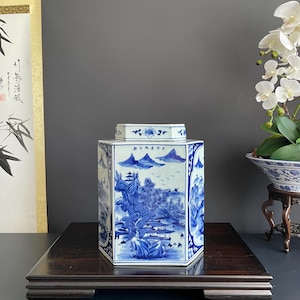 Pode incluir: Um pote de cerâmica hexagonal com uma base branca e cenas de paisagens detalhadas pintadas à mão em azul, incluindo montanhas, árvores e edifícios. A tampa também é hexagonal com um padrão floral azul correspondente e caracteres chineses.