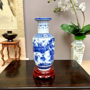 May include: A blue and white porcelain vase with a detailed landscape scene, set on a red wooden stand. The vase features a cylindrical shape with a flared top and intricate patterns. The scene depicts figures and animals. The vase is displayed on a dark wooden table.