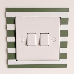 May include: A white double light switch with two rectangular toggles, set within a decorative frame. The frame has alternating stripes of white and olive green. The switch plate is mounted on a plain white wall. A modern, minimalist design.