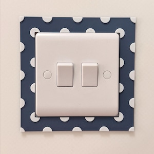 May include: A white double light switch with two rectangular toggles, set within a navy blue square plate decorated with white polka dots. The design adds a decorative touch to a standard electrical fitting.