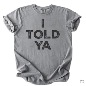 May include: Grey t-shirt featuring the text "I TOLD YA" in bold, black letters. The shirt has a crew neck and rolled-up short sleeves. The fabric looks soft and comfortable. A casual, everyday garment.