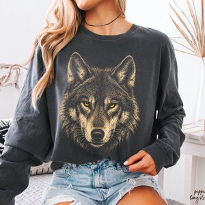 Puede incluir: Camiseta de manga larga gris oscuro con un gr&aacute;fico detallado de la cara de un lobo en dorado y amarillo. La camiseta tiene cuello redondo y un corte holgado, ideal para uso diario.