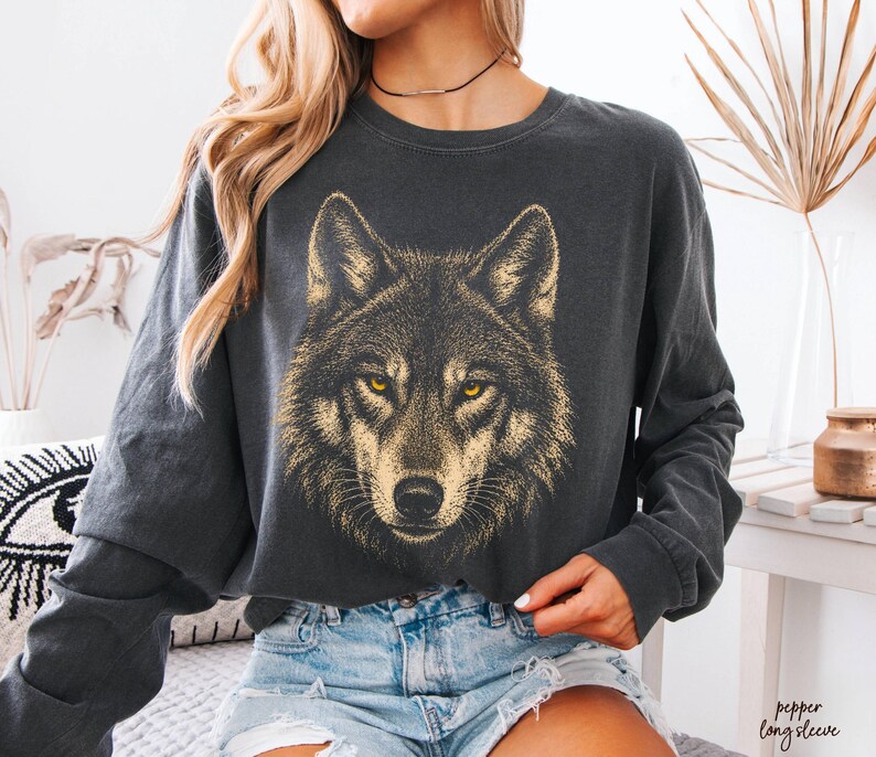 Puede incluir: Camiseta de manga larga gris oscuro con un gr&aacute;fico detallado de la cara de un lobo en dorado y amarillo. La camiseta tiene cuello redondo y un corte holgado, ideal para uso diario.