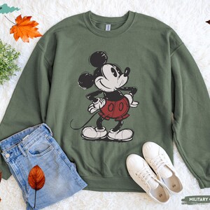 Puede incluir: Una sudadera verde oliva con un gr&aacute;fico desgastado de Mickey Mouse en negro, blanco y rojo. La sudadera se combina con vaqueros azules y zapatillas blancas. Las palabras "MILITARY GREEN" est&aacute;n impresas en la parte inferior derecha.