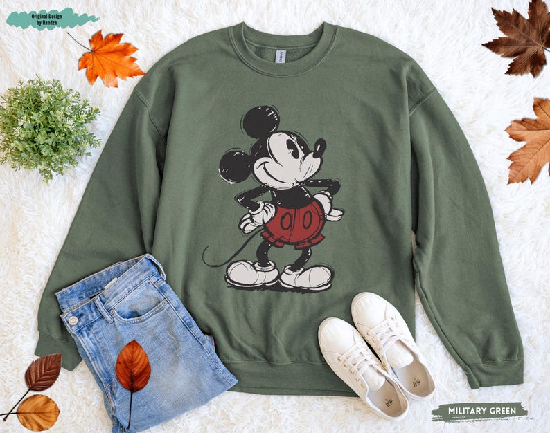 Puede incluir: Una sudadera verde oliva con un gr&aacute;fico desgastado de Mickey Mouse en negro, blanco y rojo. La sudadera se combina con vaqueros azules y zapatillas blancas. Las palabras "MILITARY GREEN" est&aacute;n impresas en la parte inferior derecha.