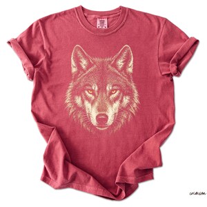 Puede incluir: Camiseta de color carmes&iacute; con una ilustraci&oacute;n detallada de la cara de un lobo. Los ojos del lobo son amarillos y el dise&ntilde;o es de un color m&aacute;s claro. La camiseta tiene mangas enrolladas y cuello redondo.