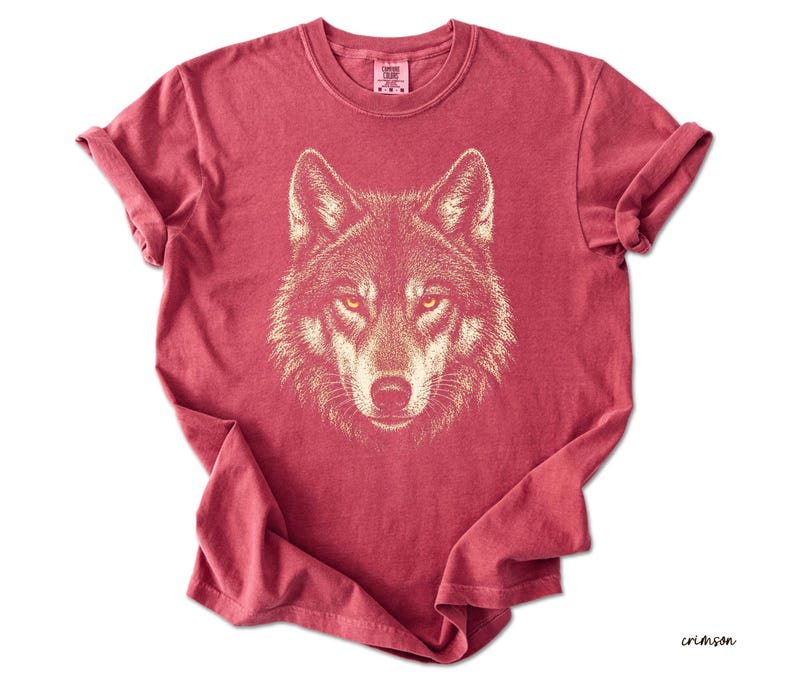 Puede incluir: Camiseta de color carmes&iacute; con una ilustraci&oacute;n detallada de la cara de un lobo. Los ojos del lobo son amarillos y el dise&ntilde;o es de un color m&aacute;s claro. La camiseta tiene mangas enrolladas y cuello redondo.