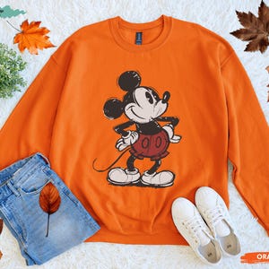 Puede incluir: Sudadera naranja con un gr&aacute;fico de Mickey Mouse en negro, blanco y rojo. La sudadera se combina con vaqueros azules y zapatillas blancas. Las palabras "Original Design by Handaz" est&aacute;n en la esquina superior izquierda. La palabra "ORANGE" est&aacute; en la esquina inferior derecha.