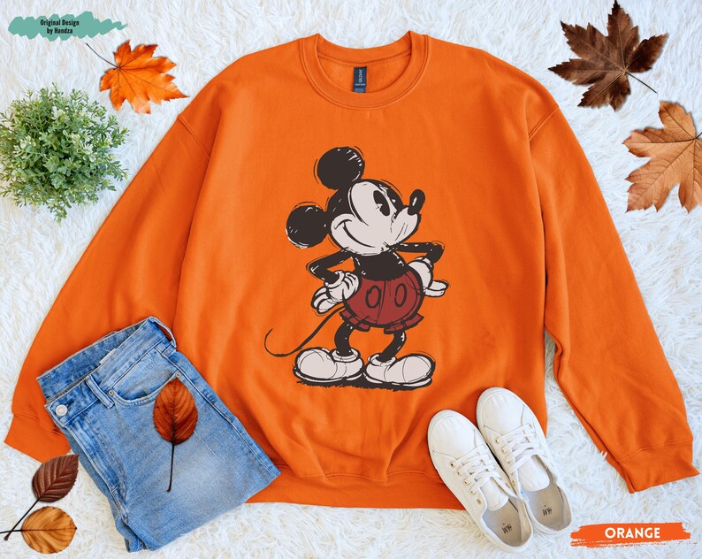 Puede incluir: Sudadera naranja con un gr&aacute;fico de Mickey Mouse en negro, blanco y rojo. La sudadera se combina con vaqueros azules y zapatillas blancas. Las palabras "Original Design by Handaz" est&aacute;n en la esquina superior izquierda. La palabra "ORANGE" est&aacute; en la esquina inferior derecha.