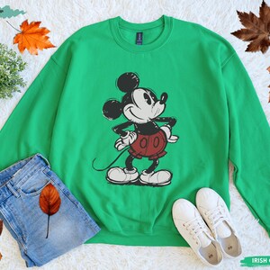 Puede incluir: Sudadera verde con un dise&ntilde;o de Mickey Mouse en negro, blanco y rojo. La sudadera se combina con vaqueros azules y zapatillas blancas. Las palabras "Irish Green" est&aacute;n impresas en la parte inferior derecha. El dise&ntilde;o es de estilo cl&aacute;sico de dibujos animados.