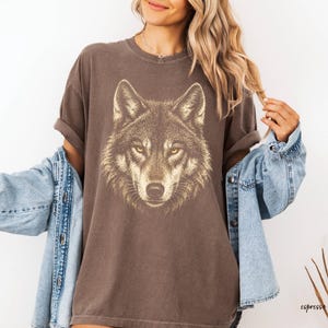 Puede incluir: Una camiseta marr&oacute;n con una ilustraci&oacute;n detallada de la cara de un lobo con ojos amarillos. La camiseta tiene un aspecto vintage y desgastado. La palabra "espresso" est&aacute; impresa en la esquina inferior derecha. Una chaqueta vaquera azul claro est&aacute; sobre la camiseta.